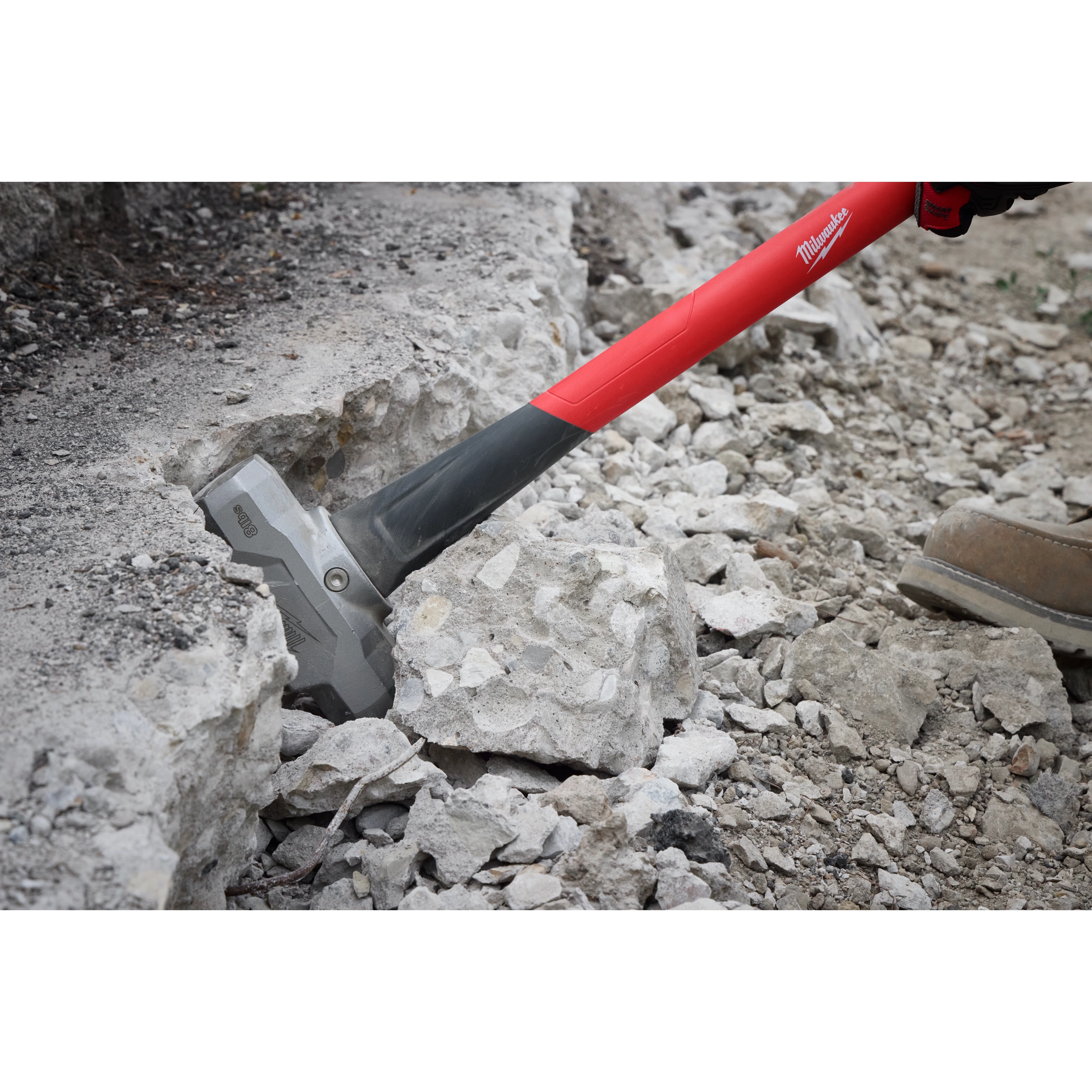 A person is using an 8lb Sledge Hammer (36" Handle) to break concrete rubble. The hammer has a bright red handle with a gray ergonomic grip. The metal head is striking the debris, showcasing its power in heavy-duty demolition tasks. Tan work boots are visible, indicating an outdoor setting.