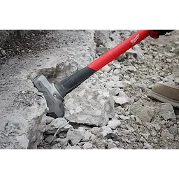 A person is using an 8lb Sledge Hammer (36" Handle) to break concrete rubble. The hammer has a bright red handle with a gray ergonomic grip. The metal head is striking the debris, showcasing its power in heavy-duty demolition tasks. Tan work boots are visible, indicating an outdoor setting.