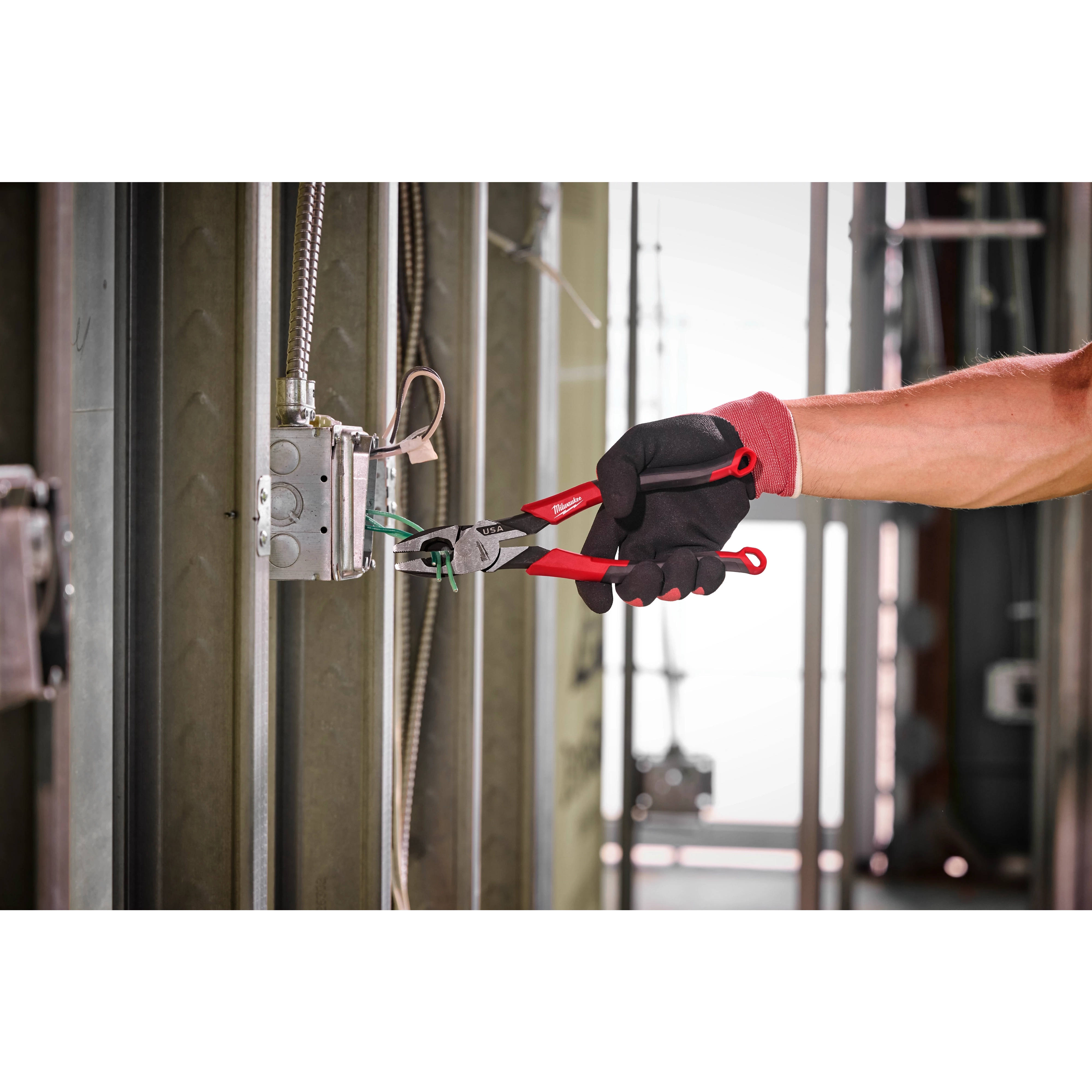 A person wearing gloves uses 9" Lineman's Comfort Grip Pliers (USA) to grip wires inside an electrical box on a metal frame.