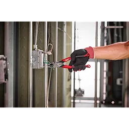 A person wearing gloves uses 9" Lineman's Comfort Grip Pliers (USA) to grip wires inside an electrical box on a metal frame.