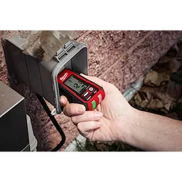 A hand is holding a Digital GFCI Receptacle Tester, inserting it into an outdoor power outlet. The device displays a reading on its digital screen, indicating its function in testing electrical outlets for safety and functionality.