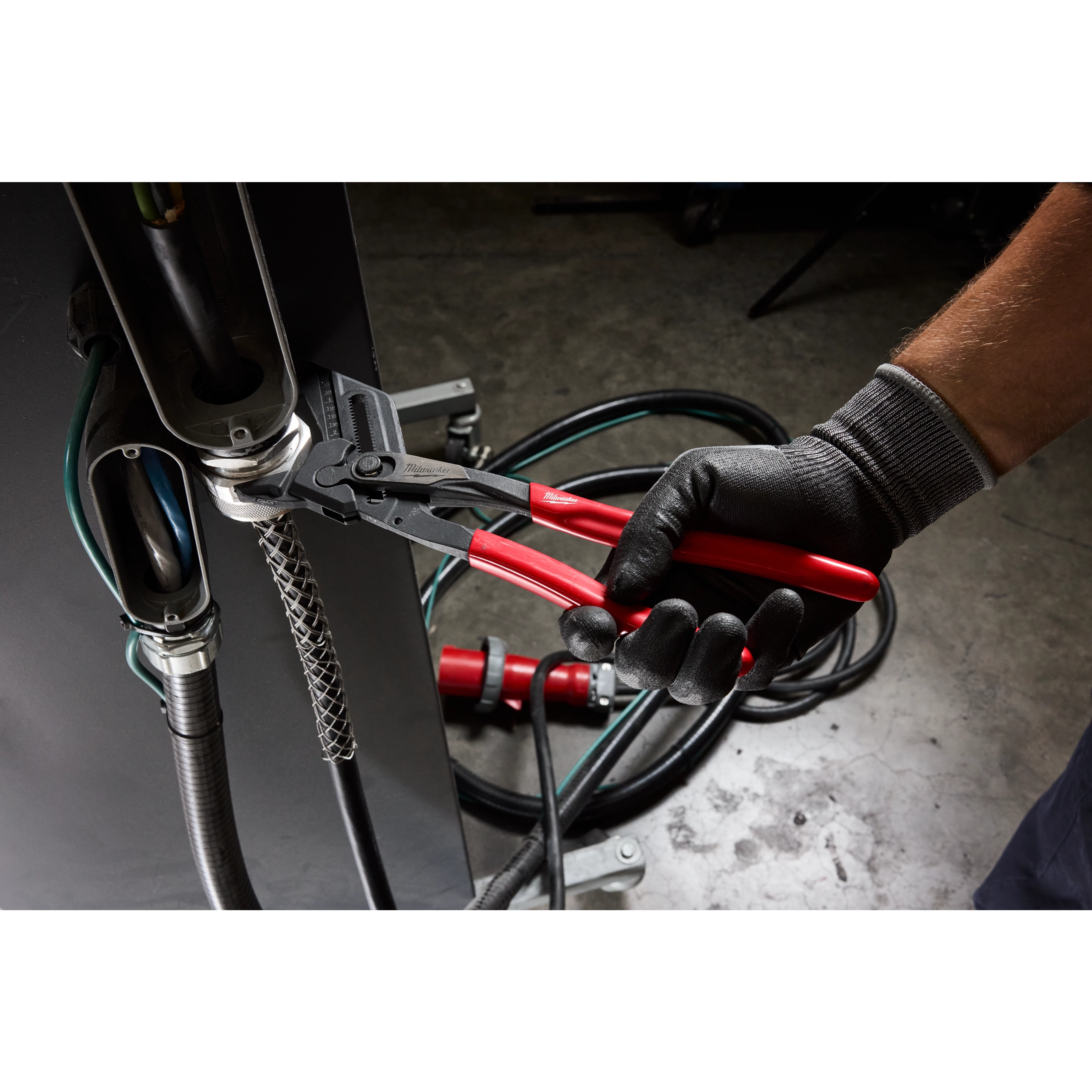 A person wearing black gloves uses a 10" Pliers Wrench to tighten a metal connector on industrial machinery. The tool has a black body and red handles, designed for gripping and turning purposes in mechanical applications.