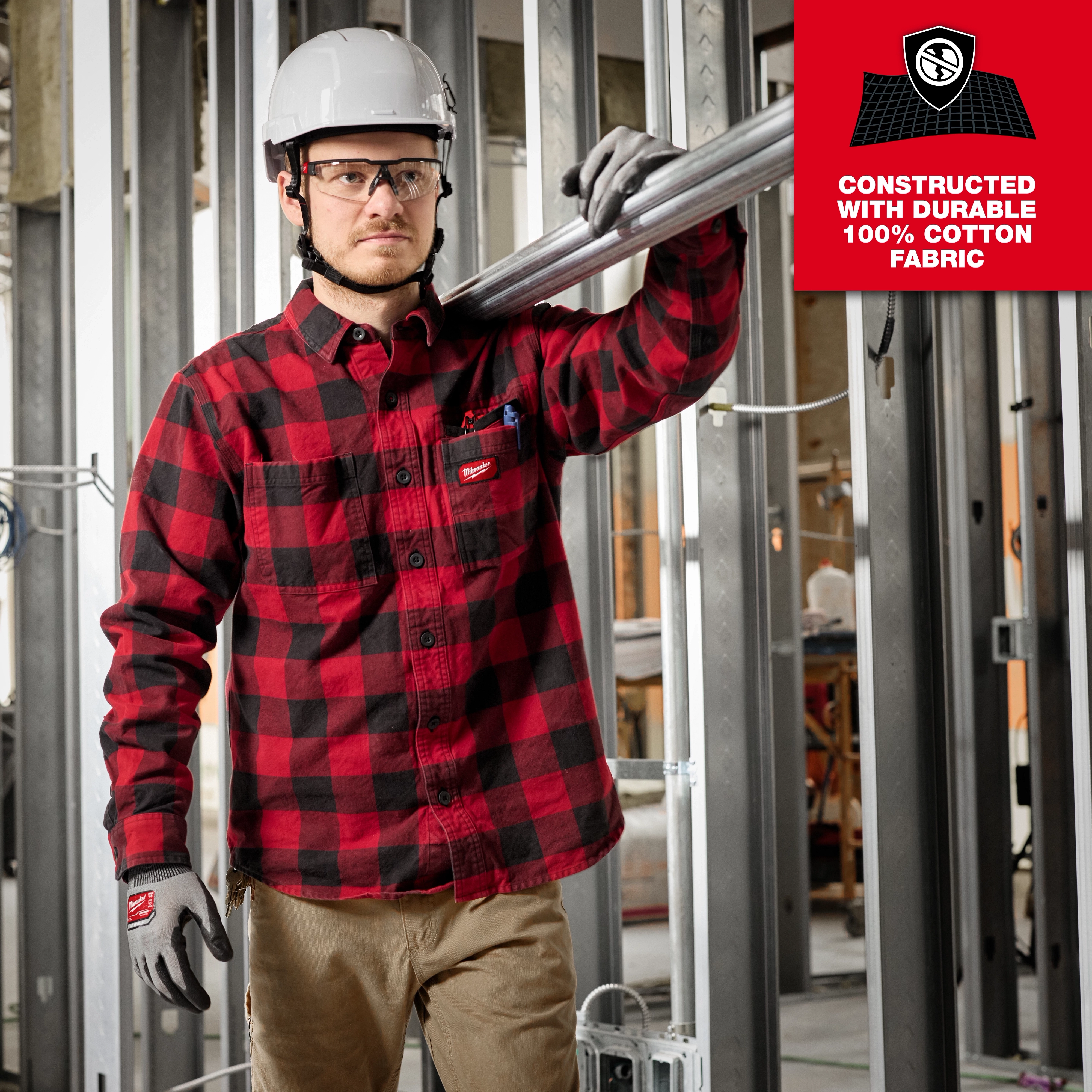 A person wearing a red and black GRIDIRON flannel shirt with a hard hat and gloves works at a construction site.