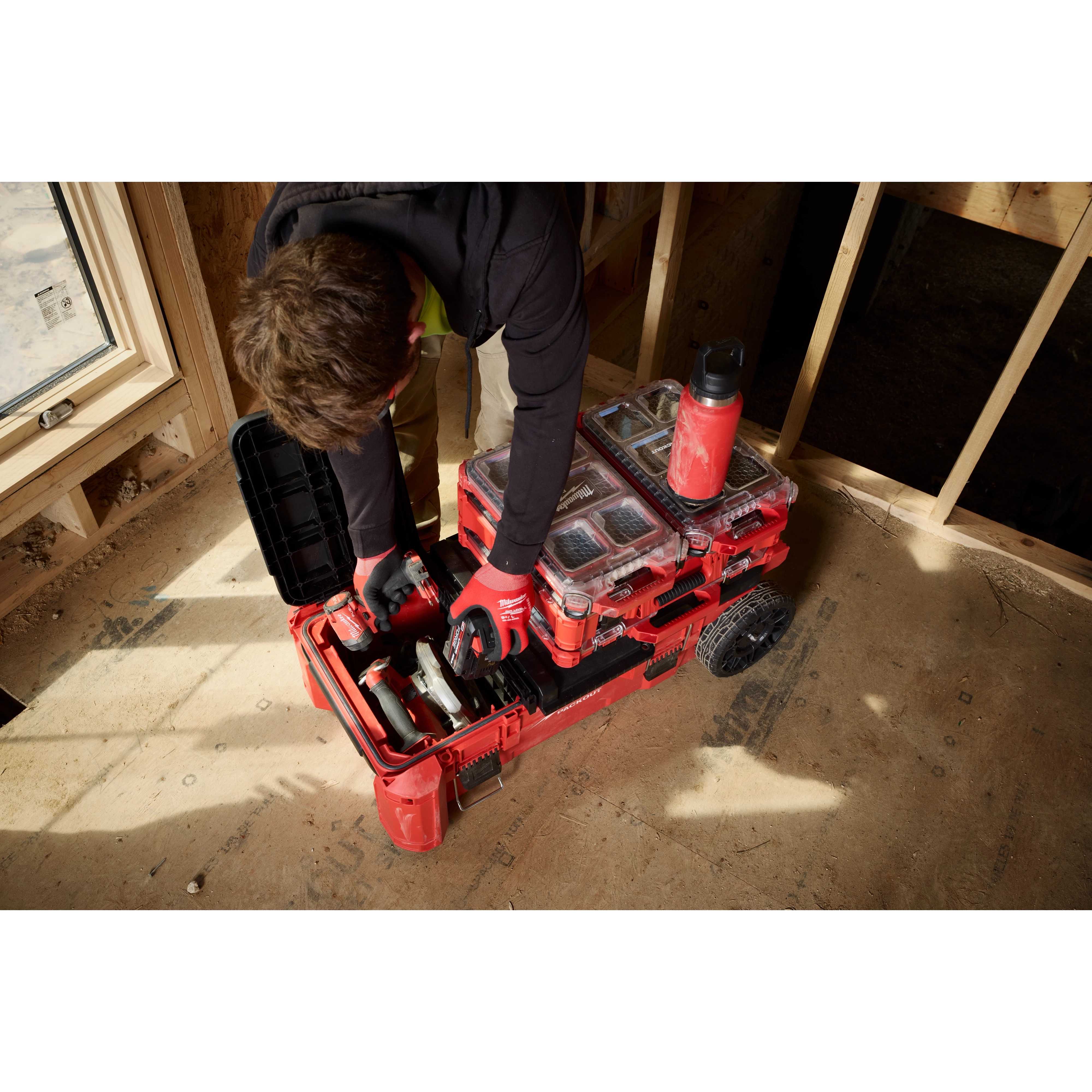 Worker organizing tools in a PACKOUT Compact Rolling Tool Chest inside a partially framed room.