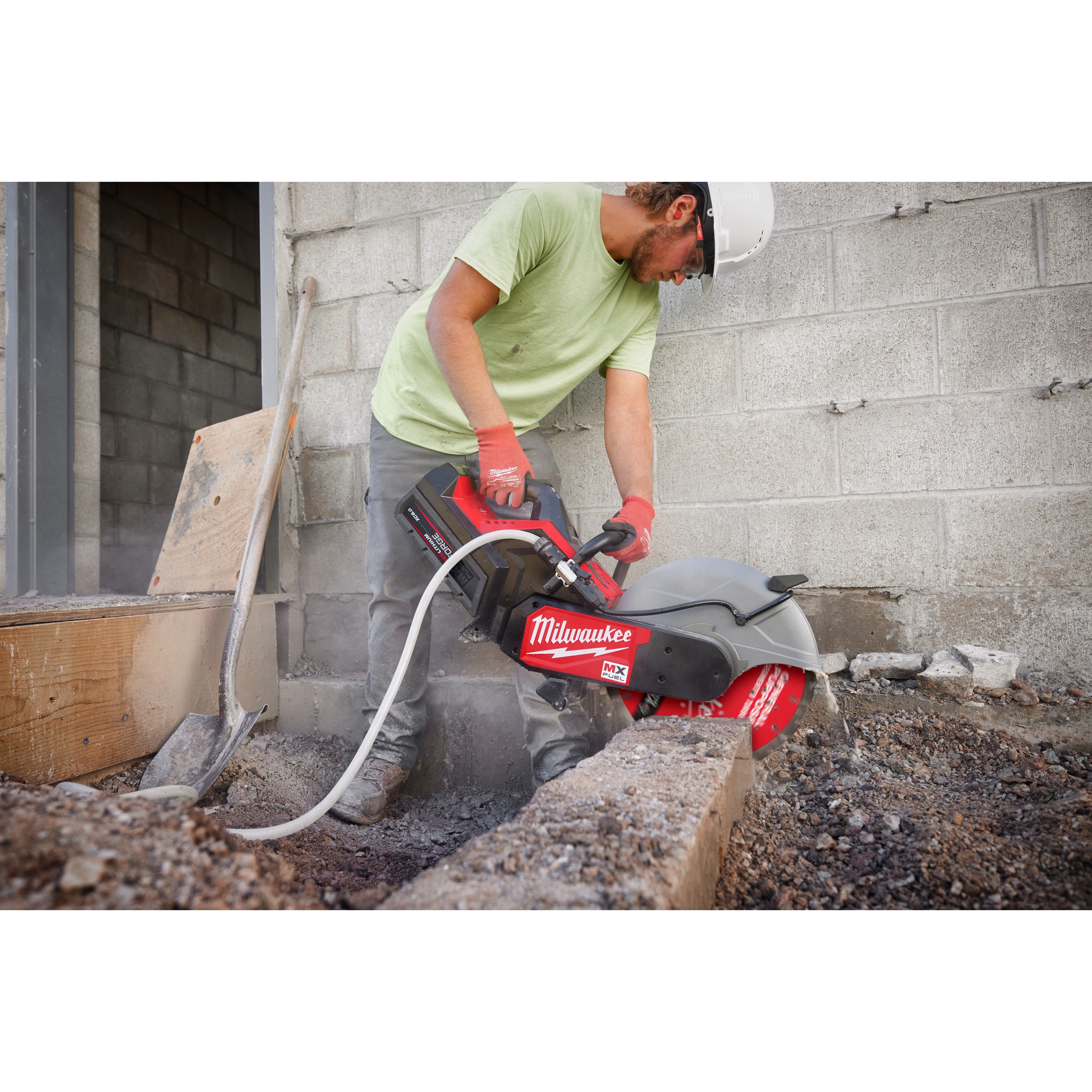 A worker is using a Milwaukee saw fitted with a 14" DIAMOND ULTRA™ Segmented Turbo, General Purpose Diamond Blade to cut through concrete at a construction site. They are wearing a green shirt, gloves, and a hard hat. The concrete wall and gravel ground are visible around the work area.