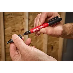 A person holds the INKZALL™ 2IN1 Deep Reach Ultra Fine Point Black Jobsite Marker in their right hand while removing the cap with their left hand. The marker is red with a black clip and tip, designed for precision marking on job sites.