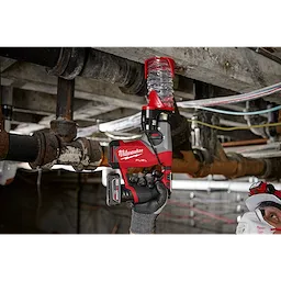 A person using the M12 FUEL™ 5/8” SDS Plus Rotary Hammer Kit is drilling into a concrete ceiling. The red and black tool is held upwards, and the person is wearing safety gear, including a helmet and mask, in an industrial setting with exposed pipes and cables.