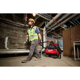 A construction worker wearing protective gear drags a red and black ROLL-ON™ 7200W/3600W 6.0kWh Power Supply through an industrial site. The power supply is mounted on a wheeled frame for easy transport. The background shows various pipes, cables, and concrete walls.