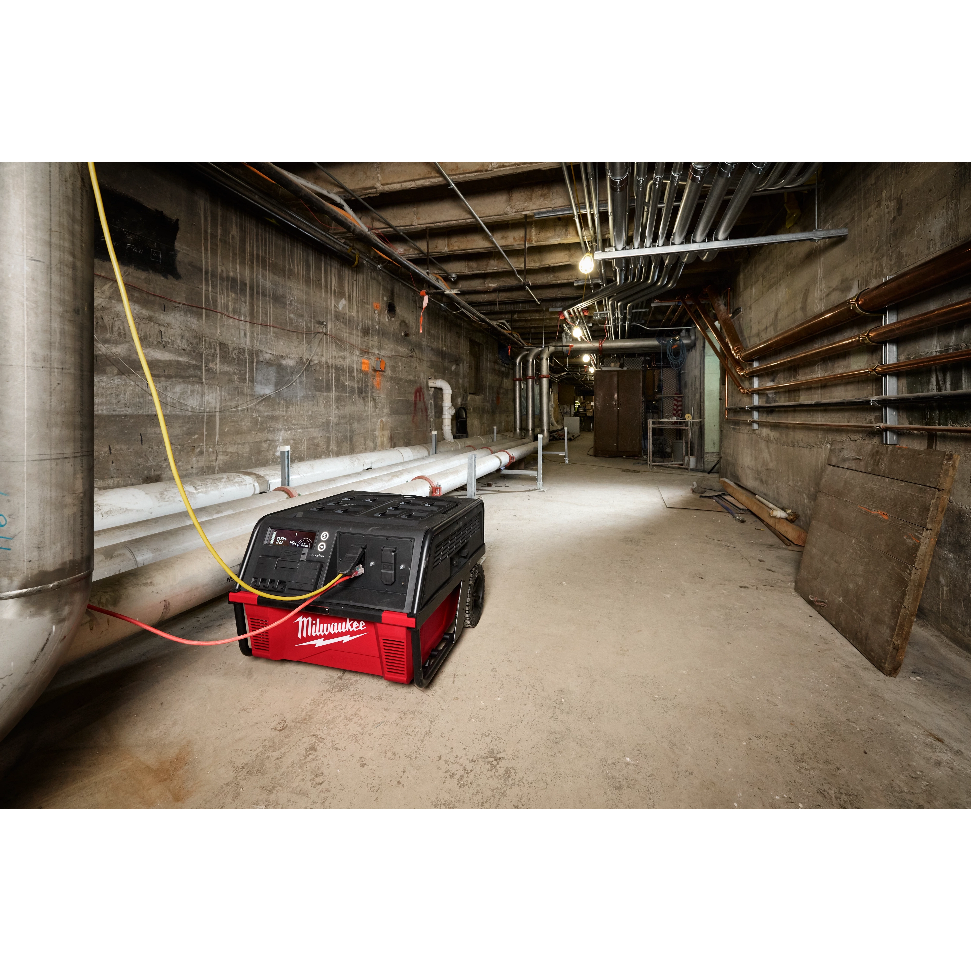 A ROLL-ON™ 7200W/3600W 6.0kWh Power Supply is positioned in an industrial basement with exposed pipes and wiring along concrete walls. The red and black unit has wheels for mobility and its cables are connected, suggesting active use. The product is designed for heavy-duty, portable power in demanding environments.