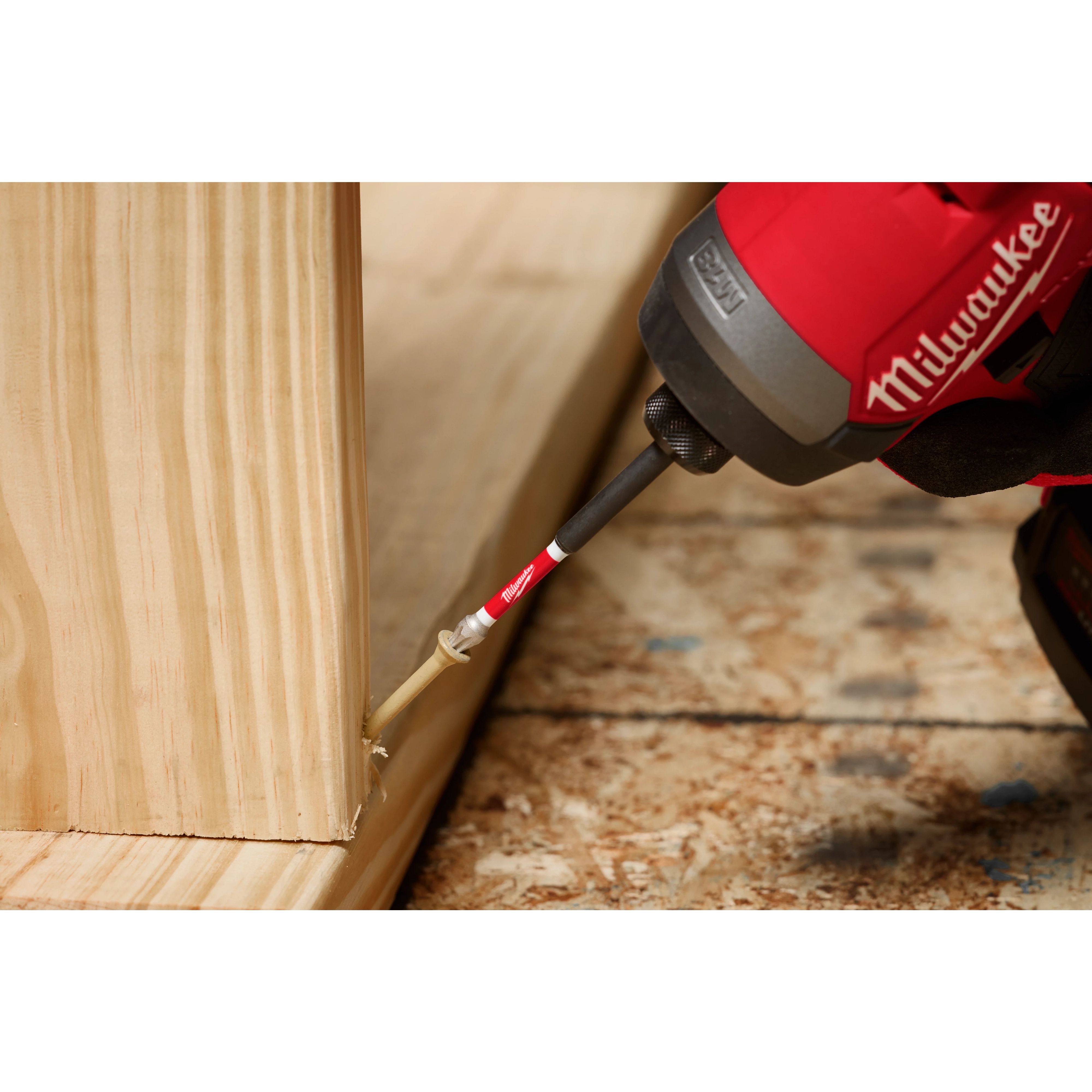 A red Milwaukee power tool with a SHOCKWAVE™ 3-1/2" Impact Torx® T25 Power Bit is driving a screw into a wooden structure. The tool is being used on a plywood surface.