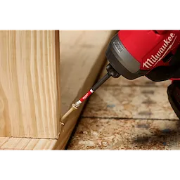A red Milwaukee power tool with a SHOCKWAVE™ 3-1/2" Impact Torx® T25 Power Bit is driving a screw into a wooden structure. The tool is being used on a plywood surface.