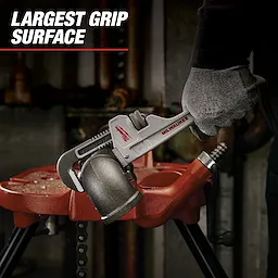 A person wearing gloves uses a Milwaukee pipe wrench to hold a pipe fitting in a workshop. The text "Largest Grip Surface" is displayed in the top left corner.