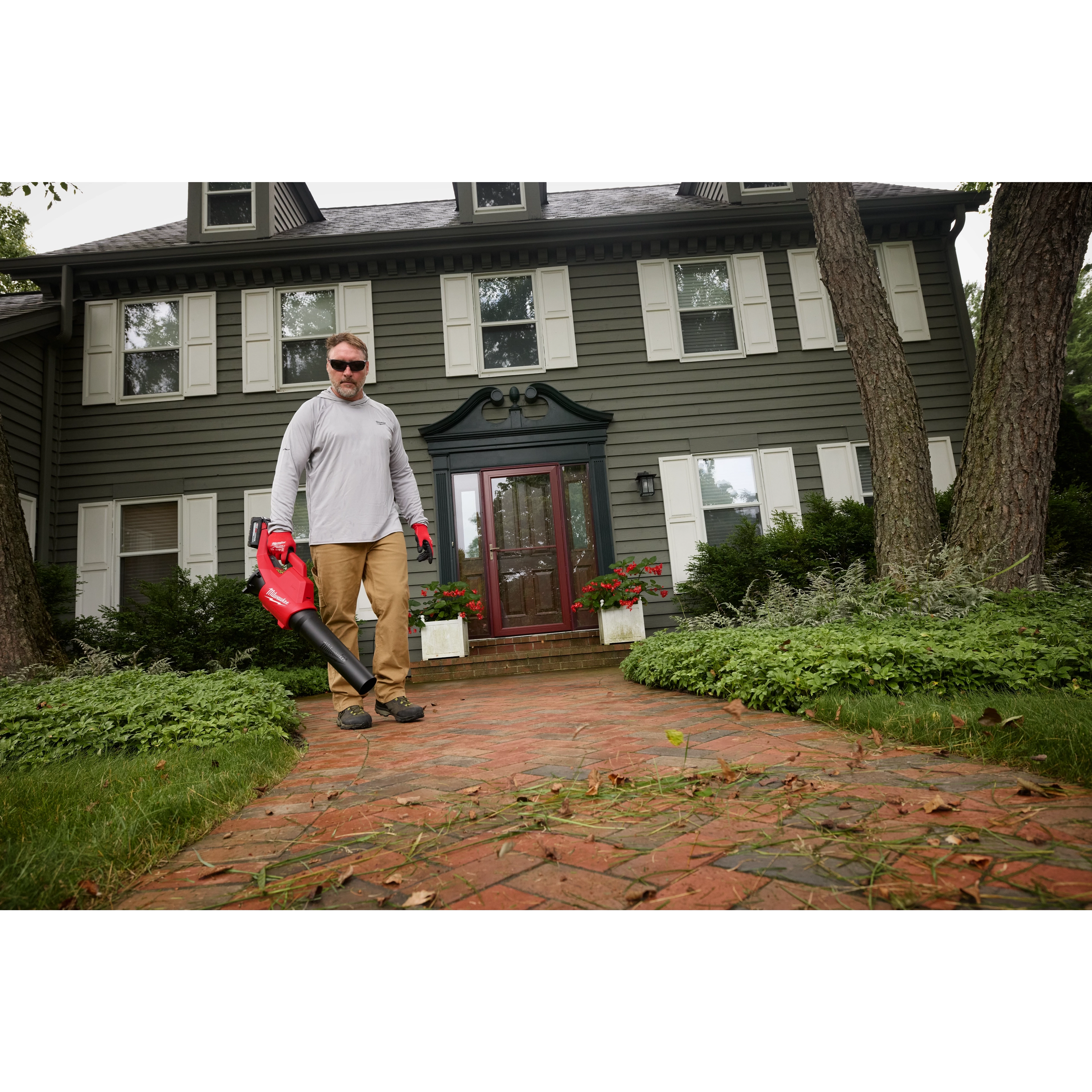 A person stands on a brick walkway in front of a green, two-story house holding an M18™ Brushless Blower. The blower is red with a black nozzle. Green bushes and trees surround the house, and fallen leaves are visible on the walkway.