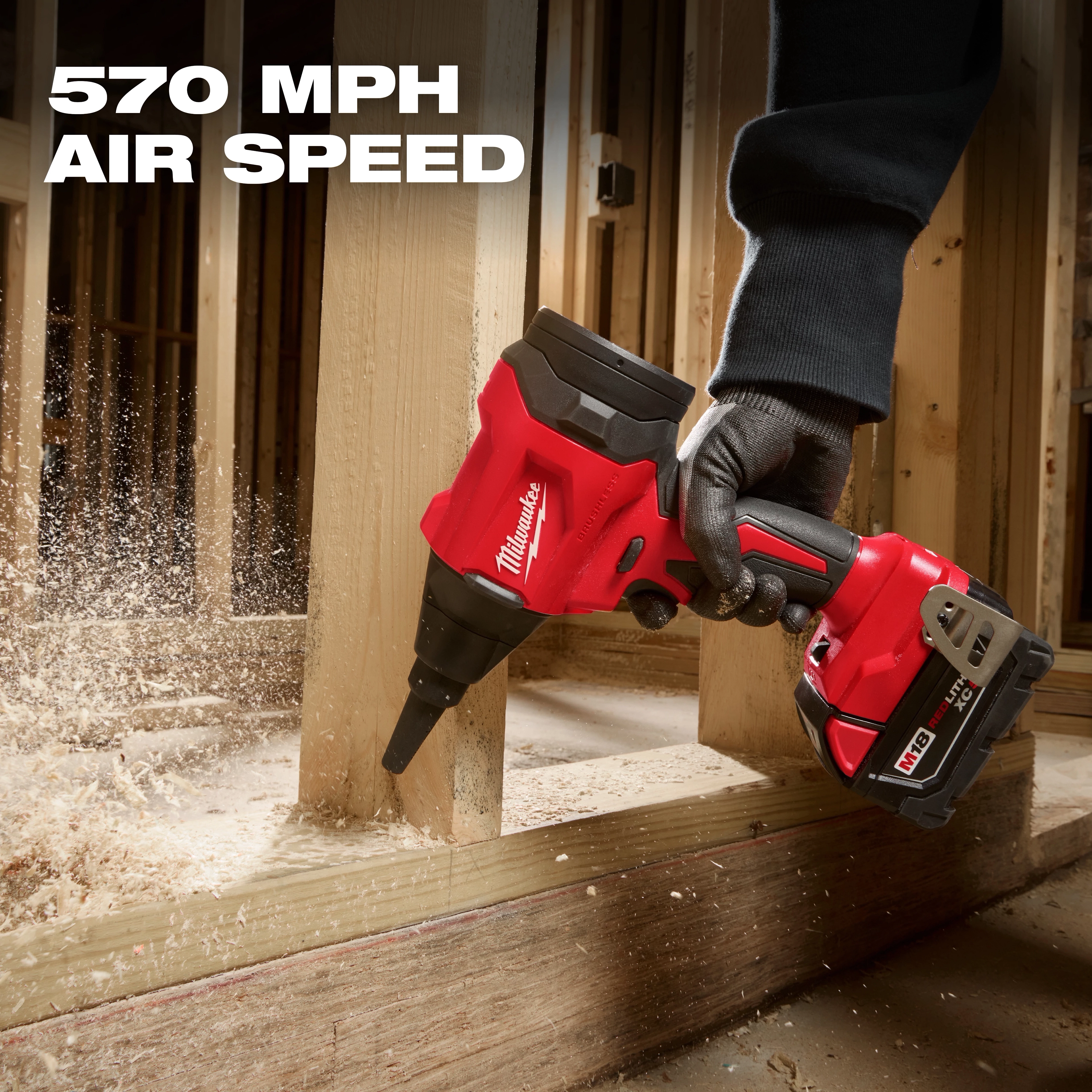 A person uses the M18™ Brushless Precision Blower by Milwaukee to clear sawdust from wood framing. The blower, which has a 570 MPH air speed, is red and black with a battery pack attached to the bottom.
