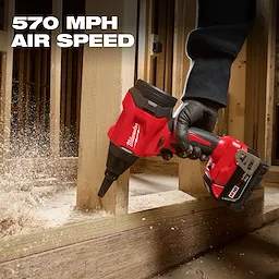 A person uses the M18™ Brushless Precision Blower by Milwaukee to clear sawdust from wood framing. The blower, which has a 570 MPH air speed, is red and black with a battery pack attached to the bottom.