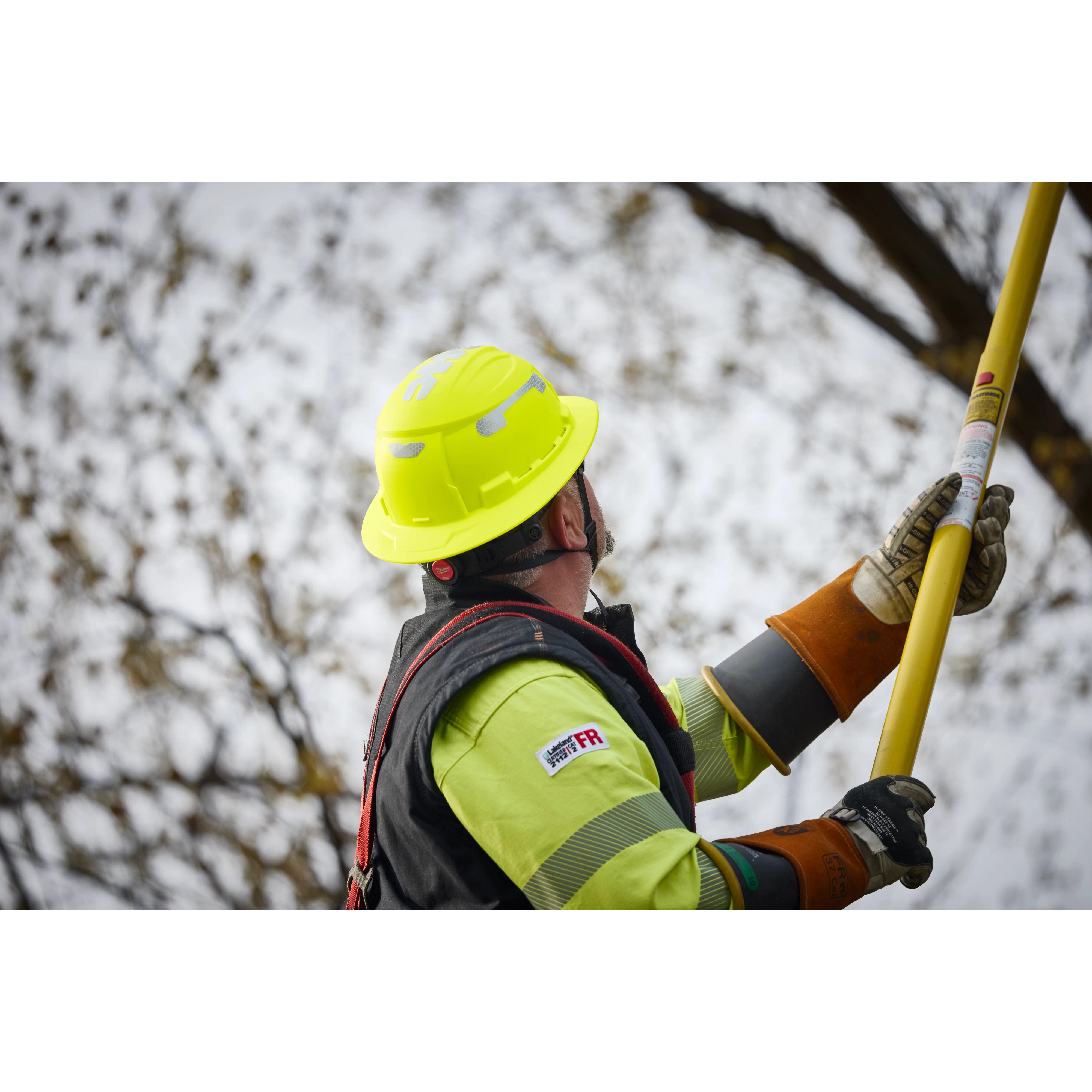 A worker wears the BOLT™ High Visibility Green Full Brim Safety Helmet – Type 2, Class E. The helmet is bright green with a full brim, and the worker holds a yellow tool while wearing protective gloves.
