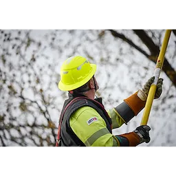 A worker wears the BOLT™ High Visibility Green Full Brim Safety Helmet – Type 2, Class E. The helmet is bright green with a full brim, and the worker holds a yellow tool while wearing protective gloves.