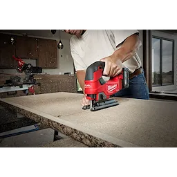 A person in a white shirt uses the M18 FUEL Jigsaw, D-Handle to cut through a piece of wood in a workshop setting.