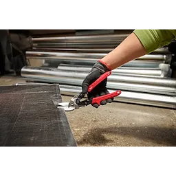 A person wearing a green sleeve and black gloves is using Left Cutting Offset Aviation Snips with red handles to cut a piece of metal sheet. Rolled metal sheets are visible in the background.