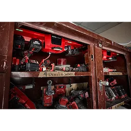 The image displays a storage cabinet containing red and black tools, with the M18™ and M12™ Gangbox Rapid Charger mounted inside the top shelf, charging multiple tool batteries. The shelves are labeled for organized storage of drills, impacts, and saws.