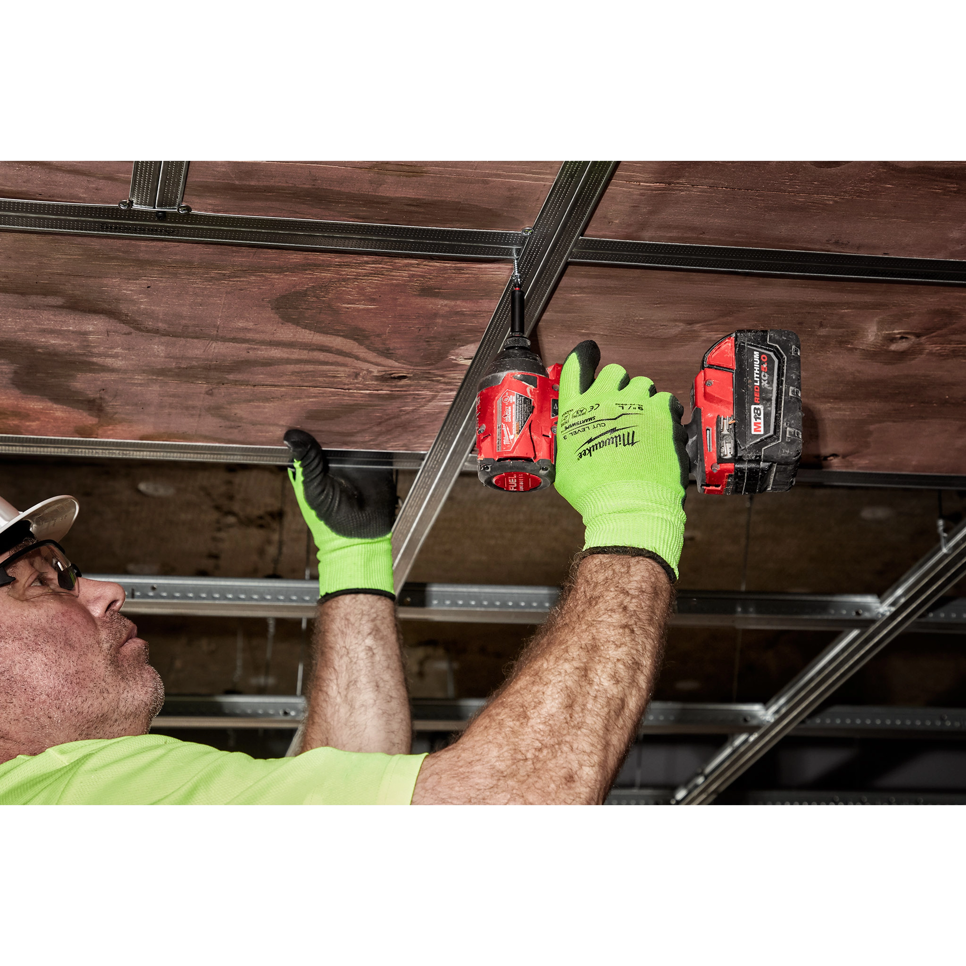 A person wearing bright green Hi Vis Cut Gloves is using a red cordless drill to secure a metal frame to a wooden ceiling.