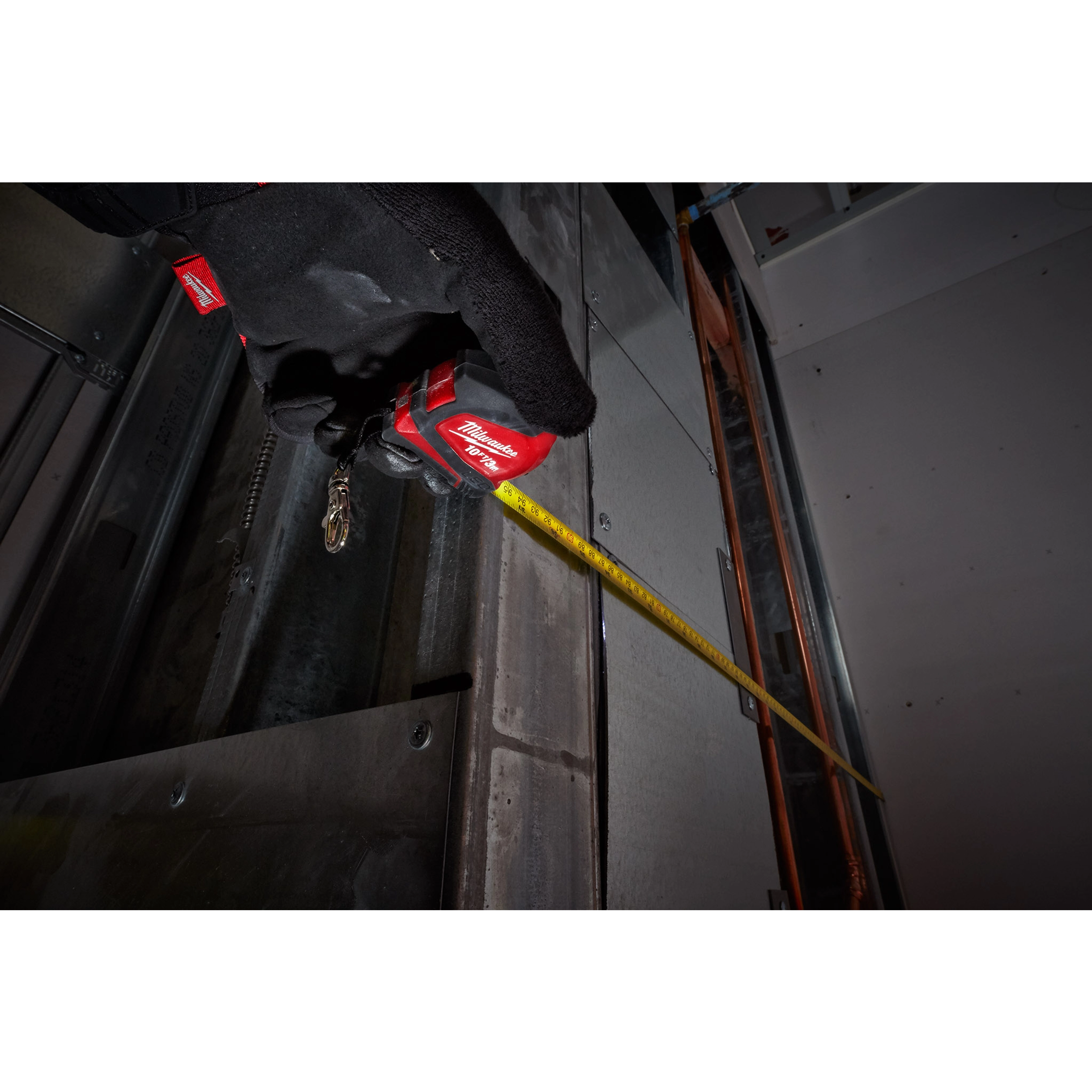 Gloved hand holding a red 10ft/3 Keychain Tape Measure with LED, extending the yellow tape while working on a metal surface.