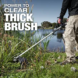 Person using an M18 FUEL QUIK-LOK Brush Cutter Attachment to trim thick brush near a lakeside.