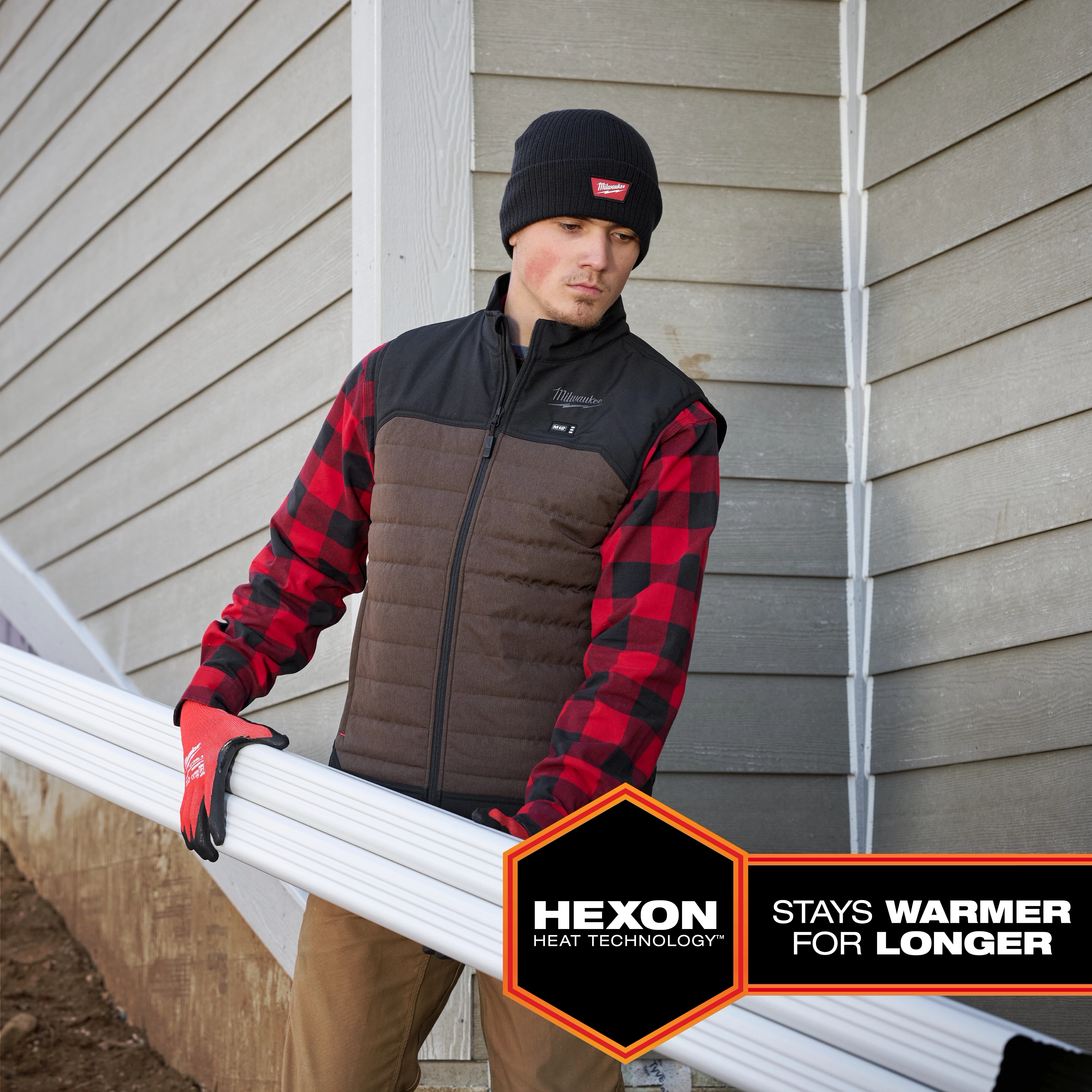 Person wearing a black and brown M12 Heated AXIS Vest with red-and-black checkered sleeves, holding white pipes. Hexon Heat Technology logo.