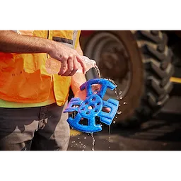 A construction worker pours water onto a blue BOLT™ Safety Helmet Cooling Liner. The worker is wearing an orange safety vest and grey pants, standing in front of heavy machinery. The liner is designed to keep helmets cool in hot conditions.