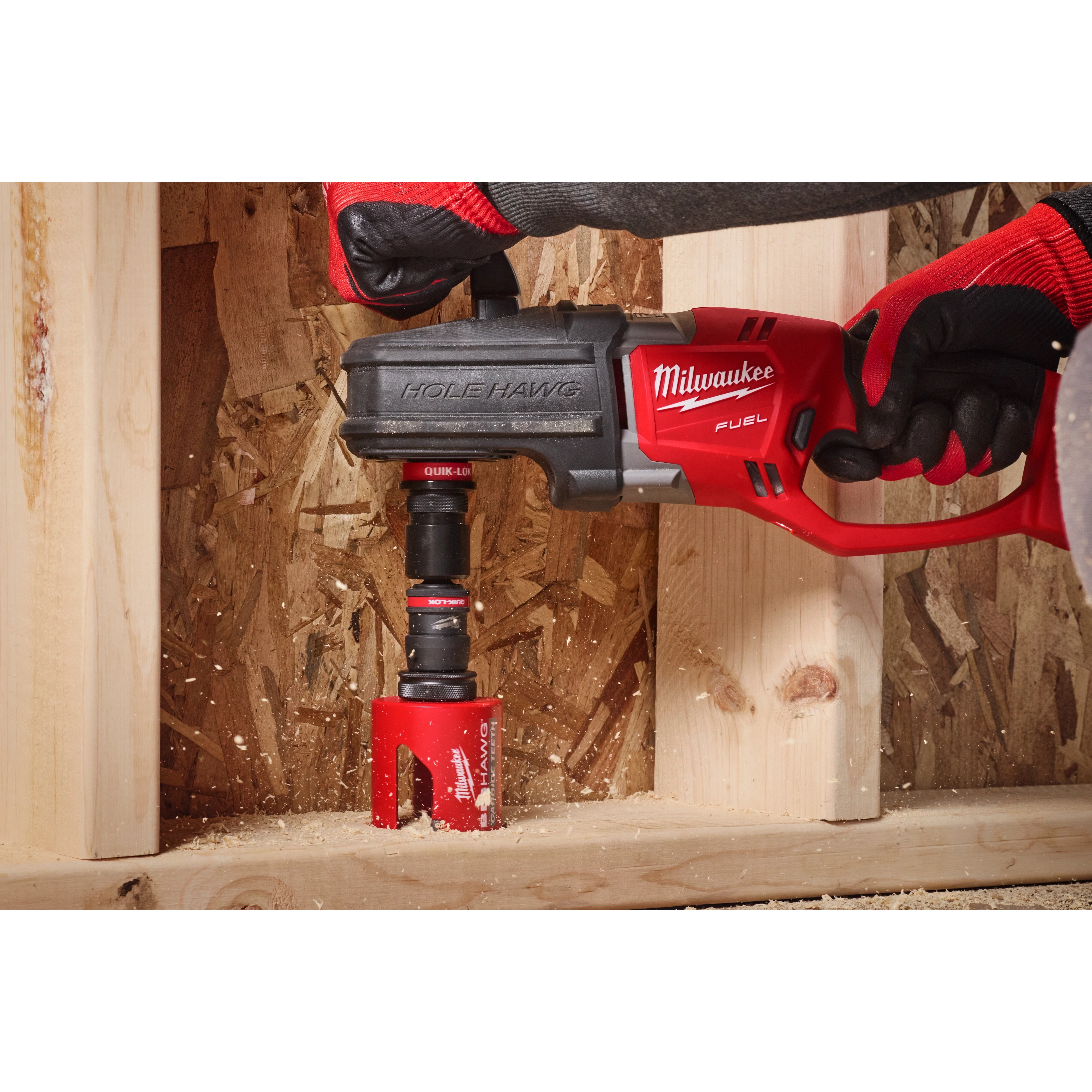 A person wearing red and black gloves uses a Hole Hawg drill with a 3/8" QUIK-LOK™ Arbor Set to drill into a wooden surface. The red drill is branded Milwaukee. Wood shavings are visible around the drilling area.