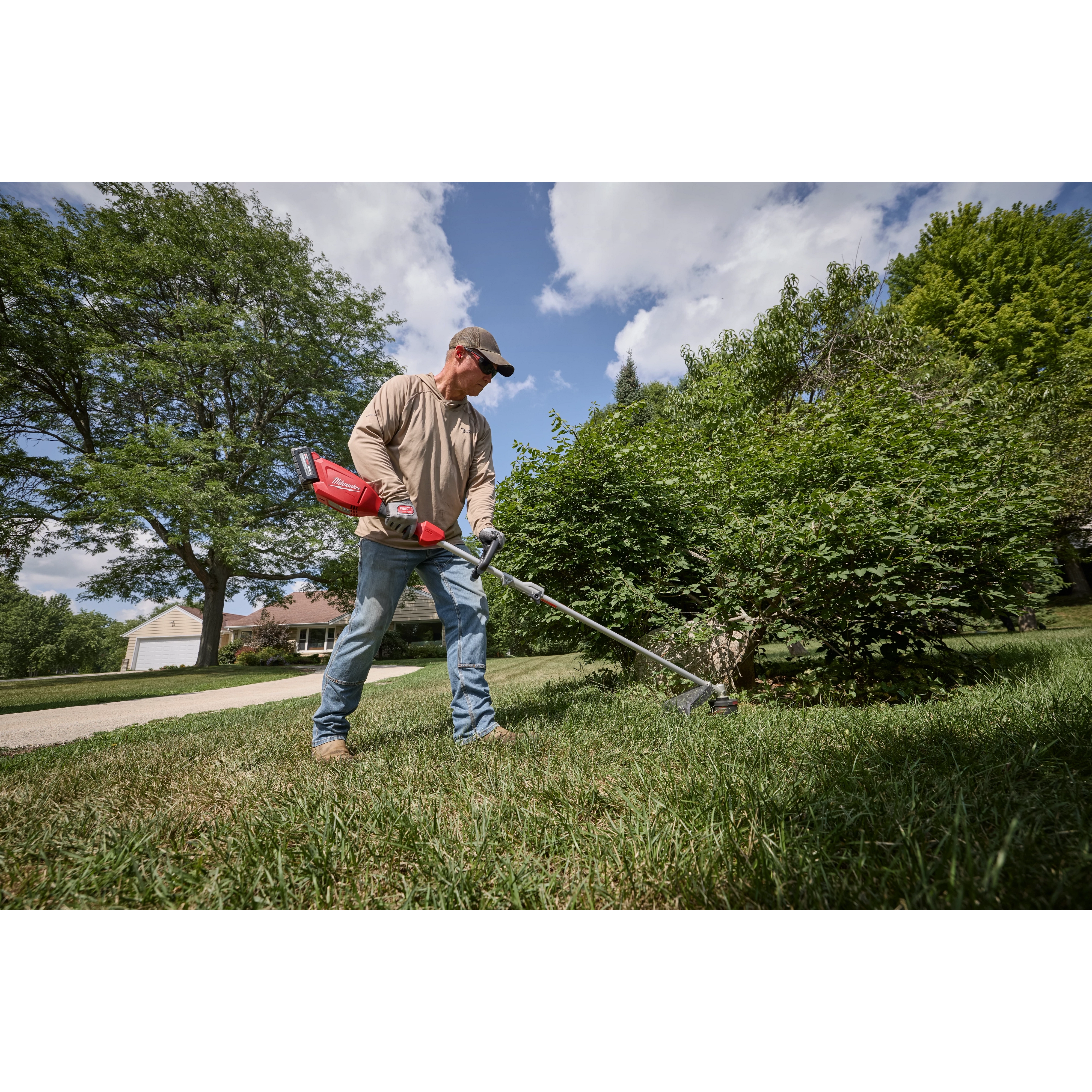 Person trimming lawn with an M18 QUIK-LOK Brushless Power Head near bushes and a house.