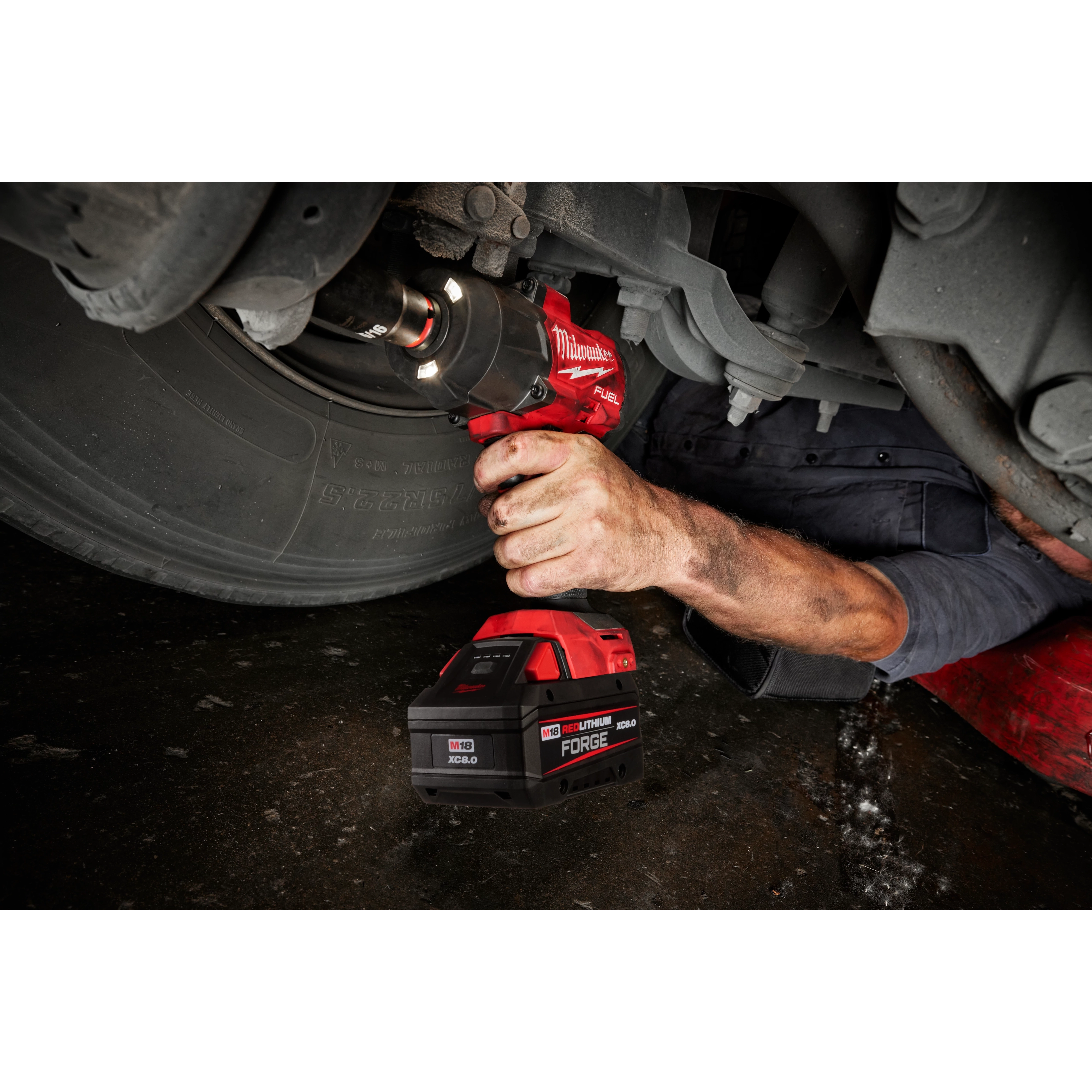 A person is using the M18™ REDLITHIUM™ FORGE™ XC6.0 battery with a power tool to work on a vehicle's wheel. The person is lying on the ground, partially under the vehicle. The battery and tool are highlighted in red and black.