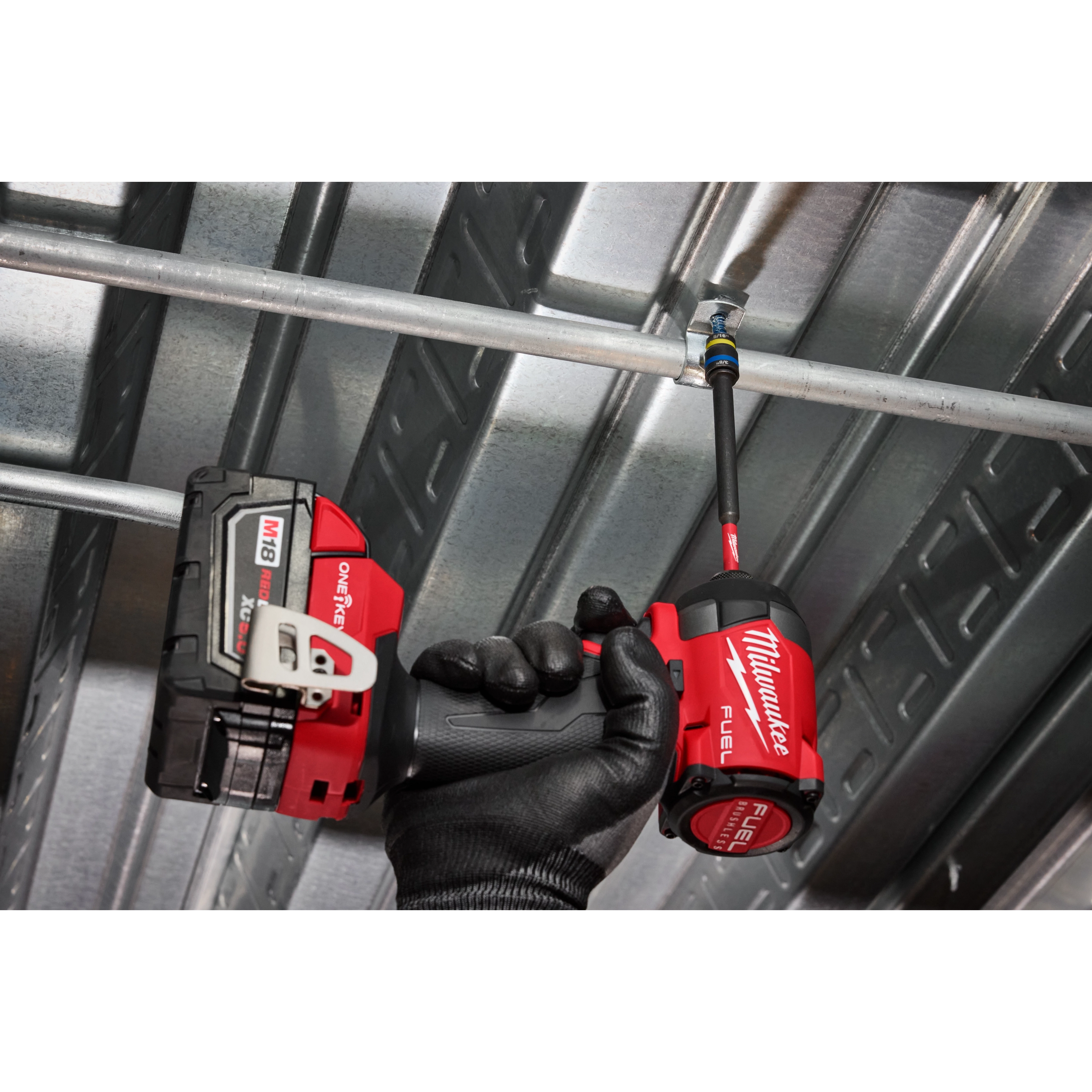 A hand wearing a black work glove holds a Milwaukee Fuel drill equipped with a SHOCKWAVE™ Impact Duty 5/16” and 3/8” x 6” QUIK-CLEAR™ 2-in-1 Magnetic Nut Driver, tightening a fastener on a metal frame. The tool and attachment are being used to secure the structure.
