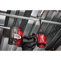 A hand wearing a black work glove holds a Milwaukee Fuel drill equipped with a SHOCKWAVE™ Impact Duty 5/16” and 3/8” x 6” QUIK-CLEAR™ 2-in-1 Magnetic Nut Driver, tightening a fastener on a metal frame. The tool and attachment are being used to secure the structure.