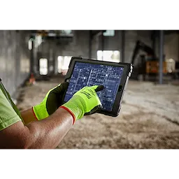 A person wearing high visibility cut level 5 polyurethane dipped gloves - L uses a tablet displaying a blueprint in a construction site.
