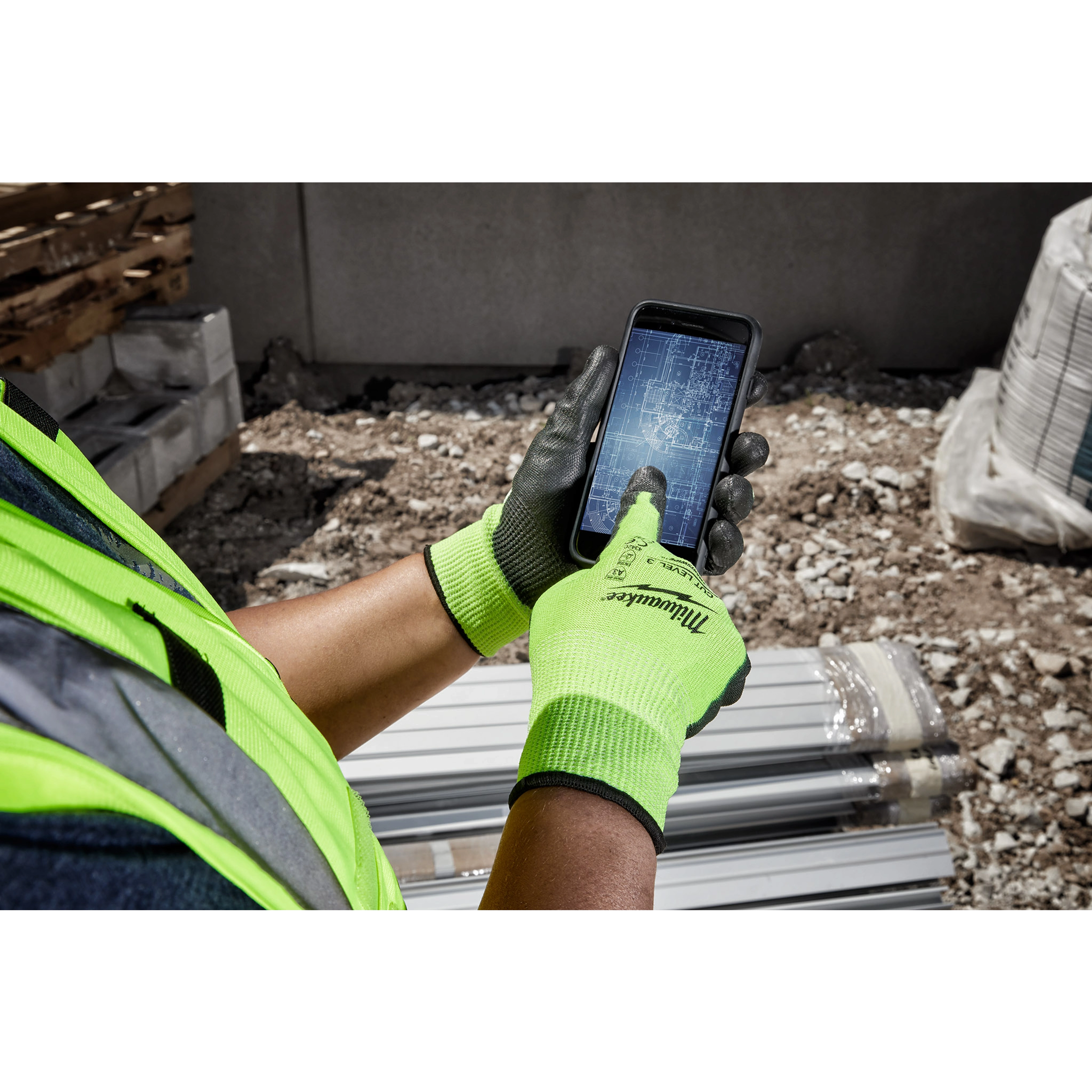 Worker wearing Hi Vis Cut Gloves uses a smartphone with blueprints on the screen at a construction site.