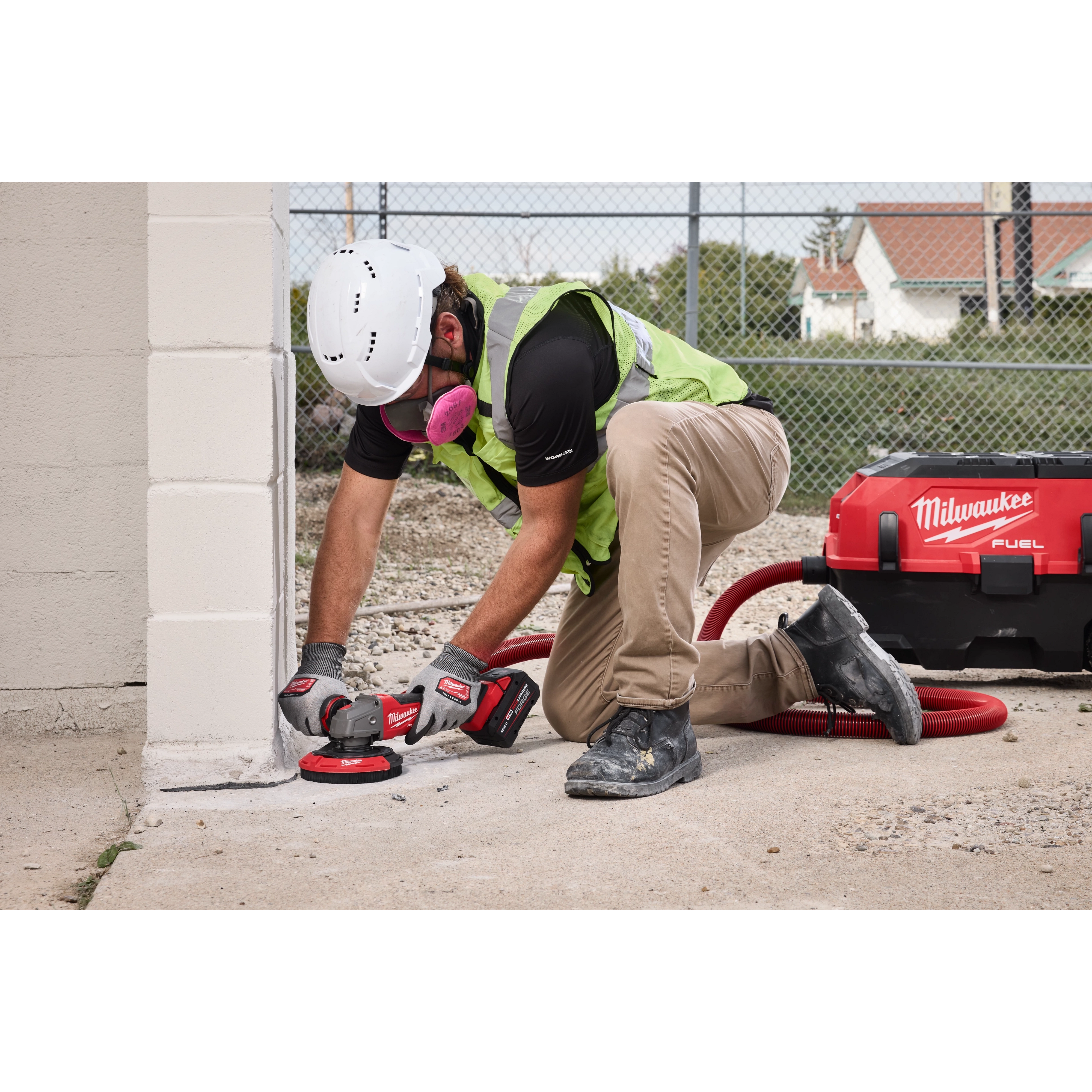 A worker in safety gear uses a Milwaukee tool equipped with the 4-1/2" - 5" Surface Grinding Dust Shroud to grind concrete near a wall. The tool is connected to a dust extractor to prevent debris inhalation and maintain a clean work area.
