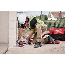A worker in safety gear uses a Milwaukee tool equipped with the 4-1/2" - 5" Surface Grinding Dust Shroud to grind concrete near a wall. The tool is connected to a dust extractor to prevent debris inhalation and maintain a clean work area.