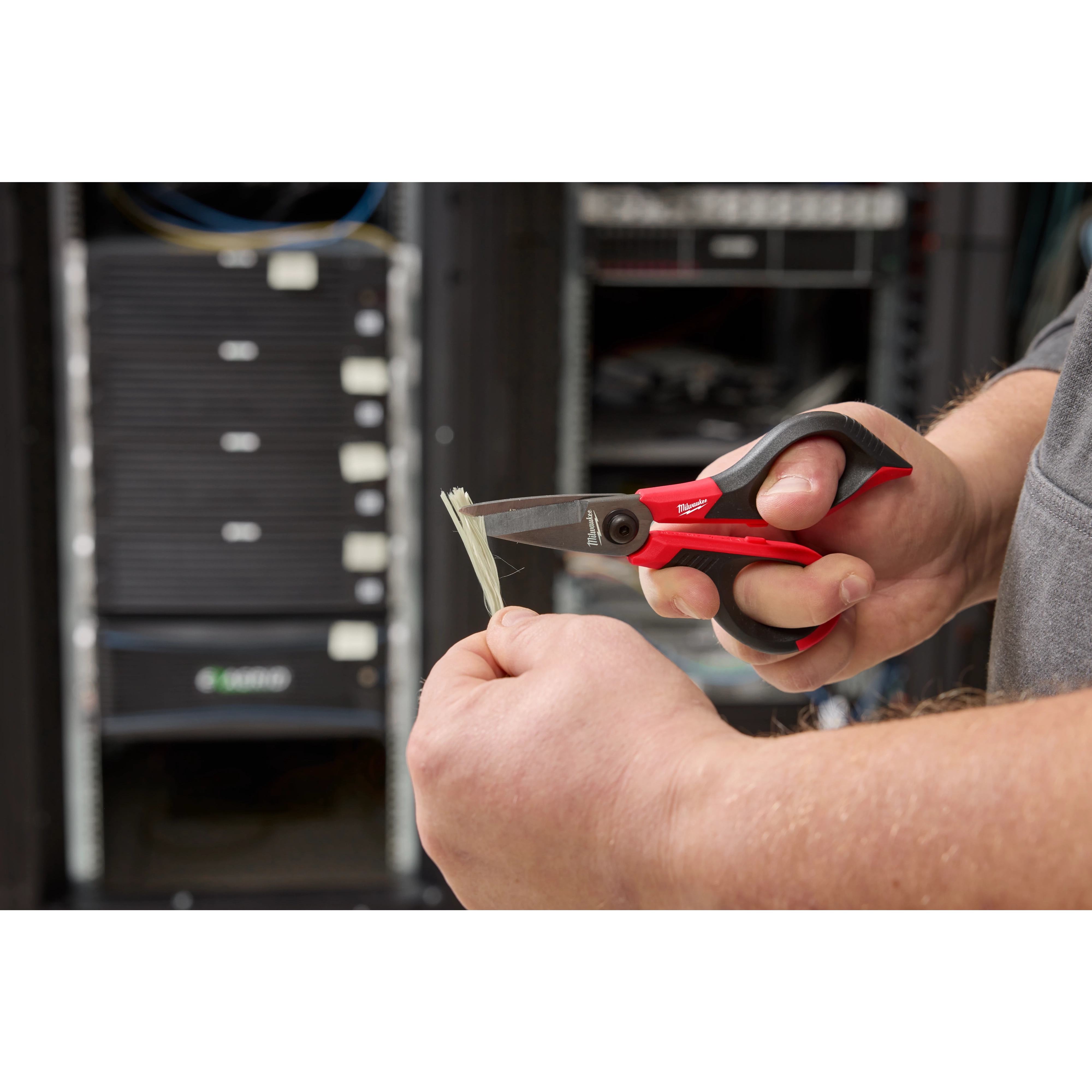 A person uses Kevlar® Shears with red and black handles to cut a Kevlar fiber in front of server equipment in a data center.