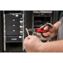 A person uses Kevlar® Shears with red and black handles to cut a Kevlar fiber in front of server equipment in a data center.