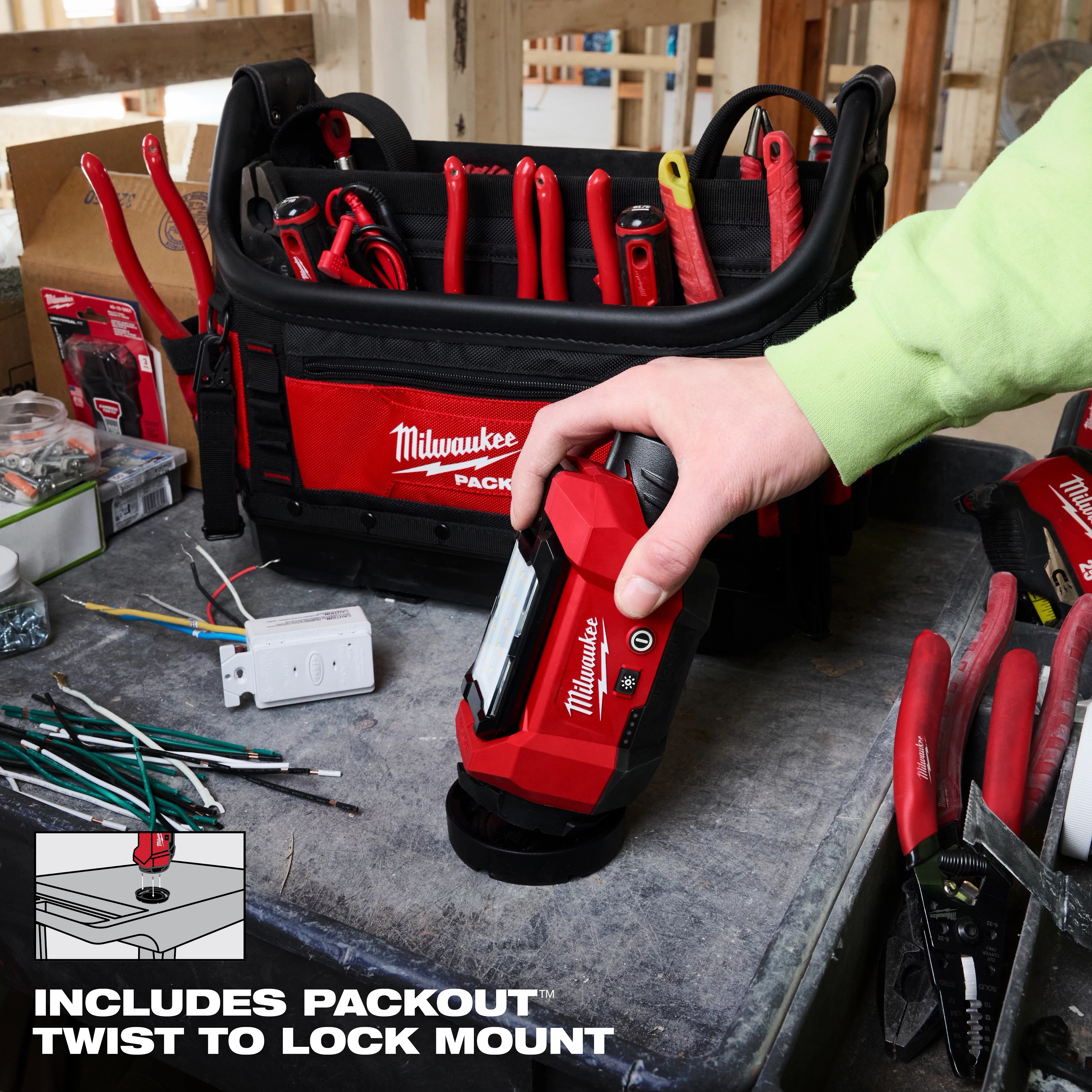A person holds the Milwaukee M12™ ROVER™ Multi-Directional Flood Light with PACKOUT™ Compatibility. The light is placed on a workbench next to various tools and a Milwaukee tool bag. The image highlights the PACKOUT™ Twist to Lock Mount feature.