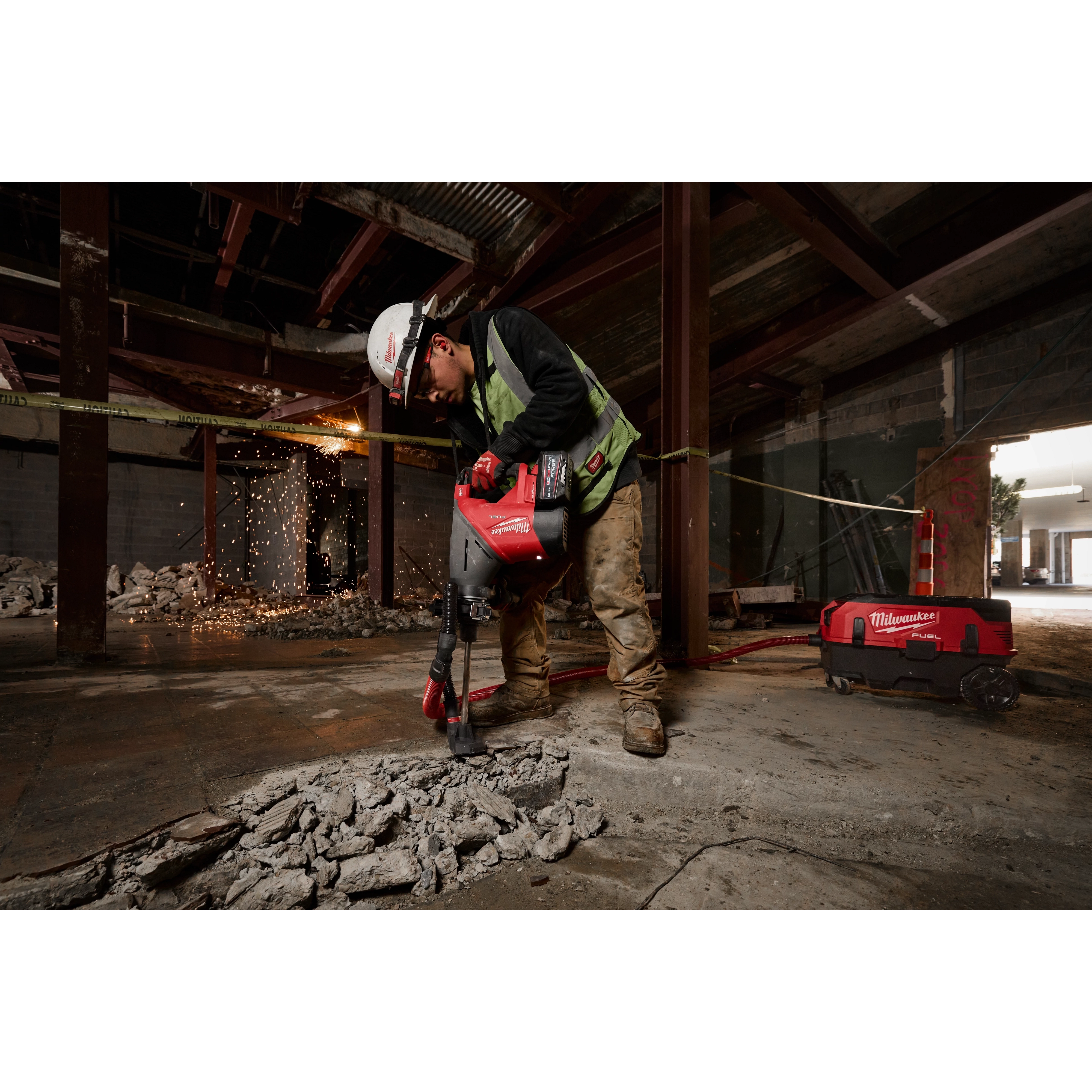 A construction worker in safety gear uses the M18 FUEL(tm) 1-3/4" SDS MAX Rotary Hammer Kit w/ONE-KEY to break concrete in a partially demolished building. Sparking debris and a red toolbox are visible in the background.