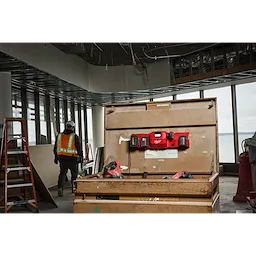 The M18™ and M12™ Gangbox Rapid Charger is mounted inside a large, open wooden toolbox on a construction site. The background shows a worker in a safety vest and helmet, with unfinished walls and large windows overlooking a body of water.
