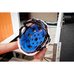 A pair of hands holds a white helmet featuring the BOLT™ Safety Helmet Cooling Sweat Band and Liner, along with the BOLT™ Safety Helmet Cooling Liner in blue, designed for enhanced comfort and cooling. The background shows outdoor steps and part of an orange and white building.