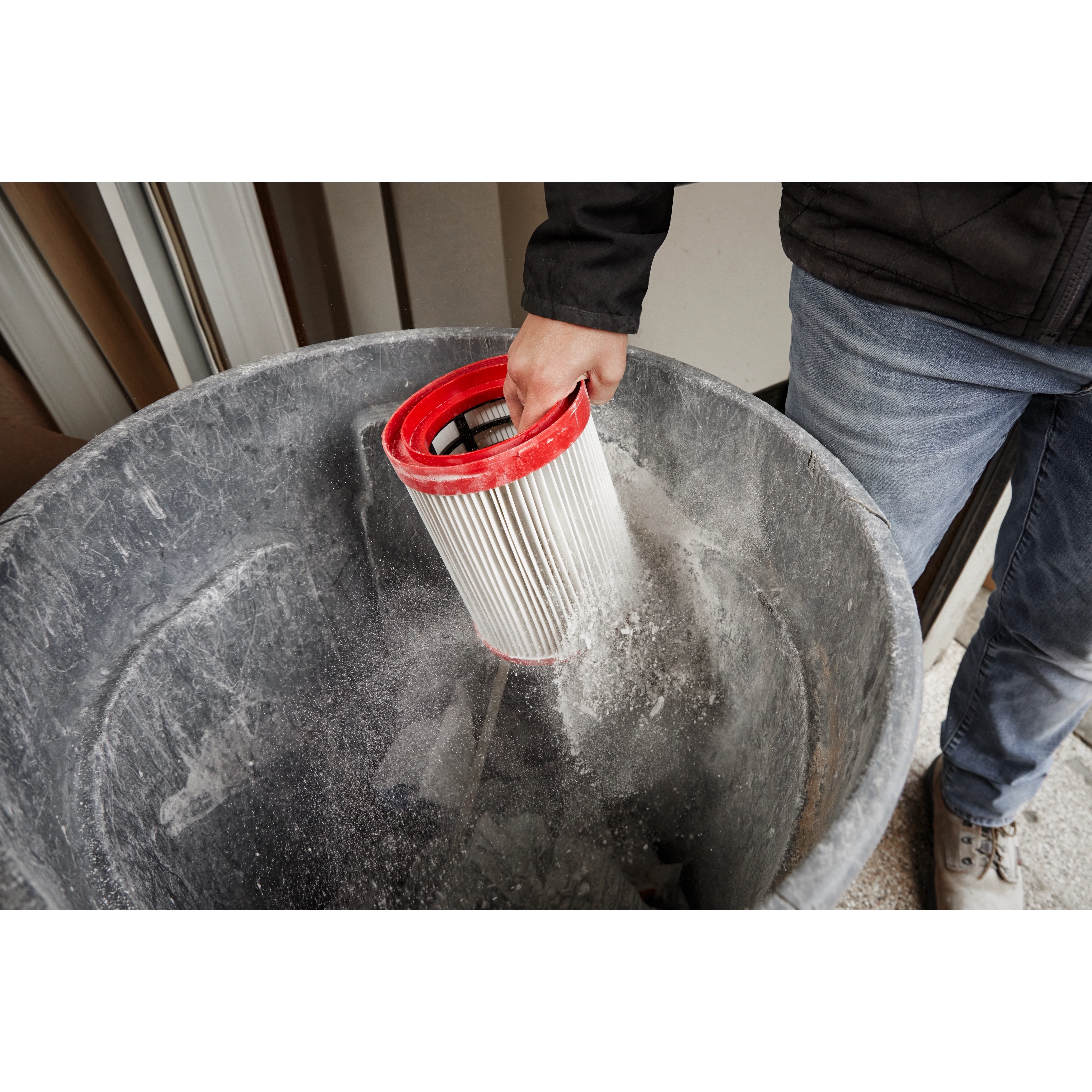 Person cleaning a Milwaukee Large Wet/Dry Vacuum HEPA Filter by tapping it over a trash can in a workshop.
