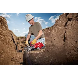 A person kneels in a trench using the FORCELOGIC™ M18™ 3" Underground Cable Cutter Kit to cut a cable. The red cable cutter is held with both hands and is cutting through a thick underground cable. The kit appears durable and suitable for heavy-duty tasks.