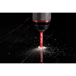 A close-up of a Milwaukee® 1/4" DIAMOND MAX™ Hole Saw Bit being used with a drill on a black surface, producing dust as it drills.