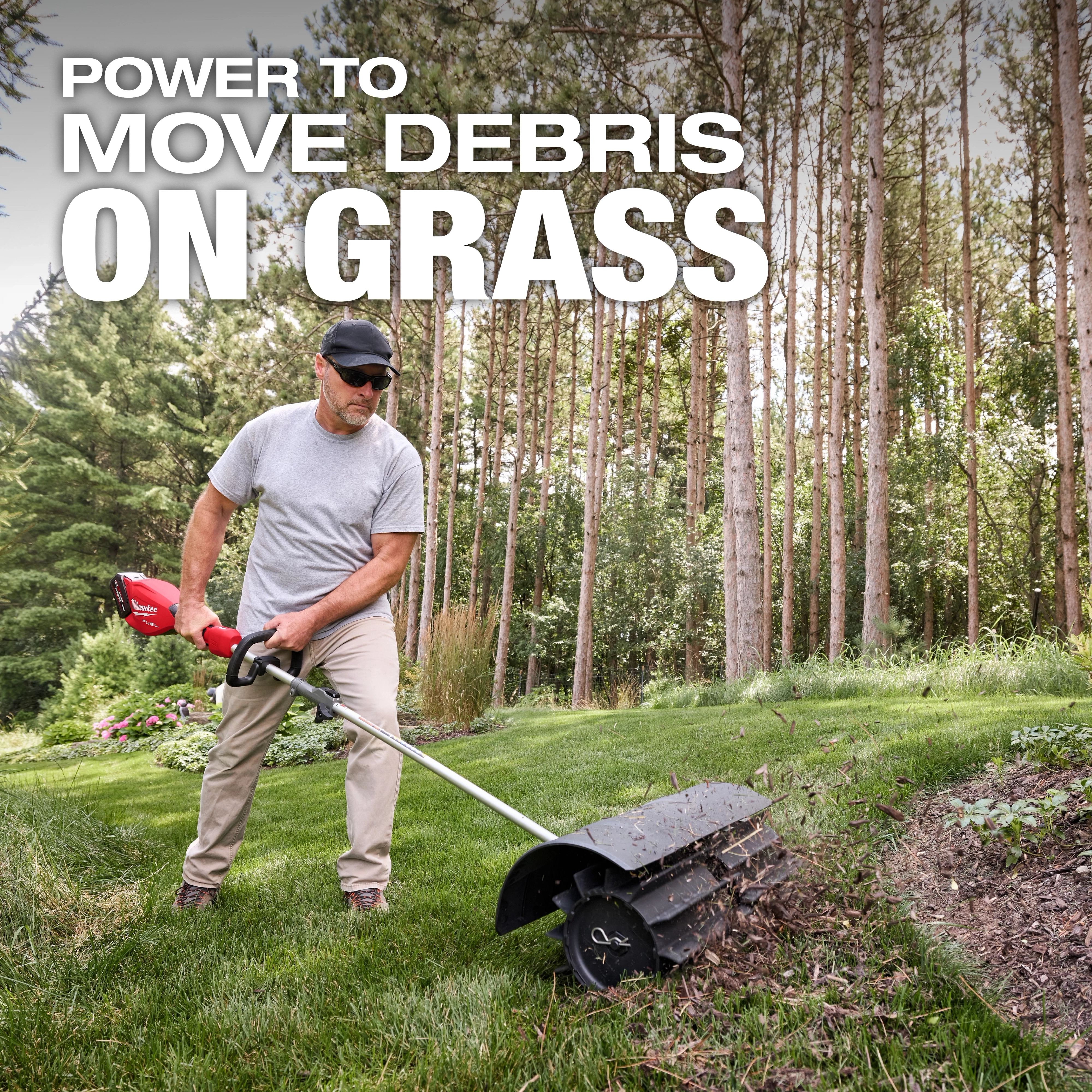 Person using an M18 FUEL QUIK-LOK Rubber Broom Attachment to clear debris from grass near a wooded area.