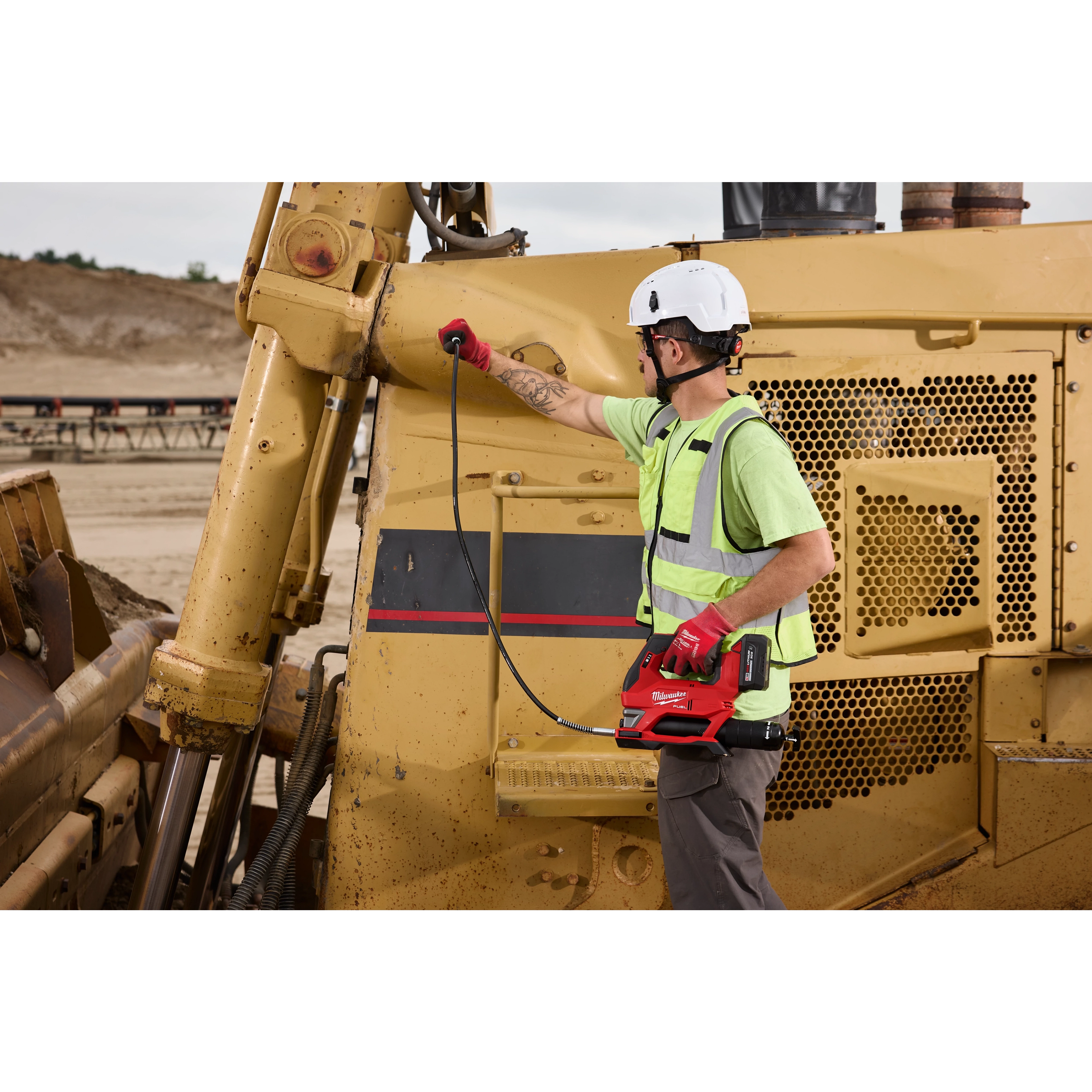 Worker using an M18 FUEL Grease Gun to service a large construction vehicle outdoors.