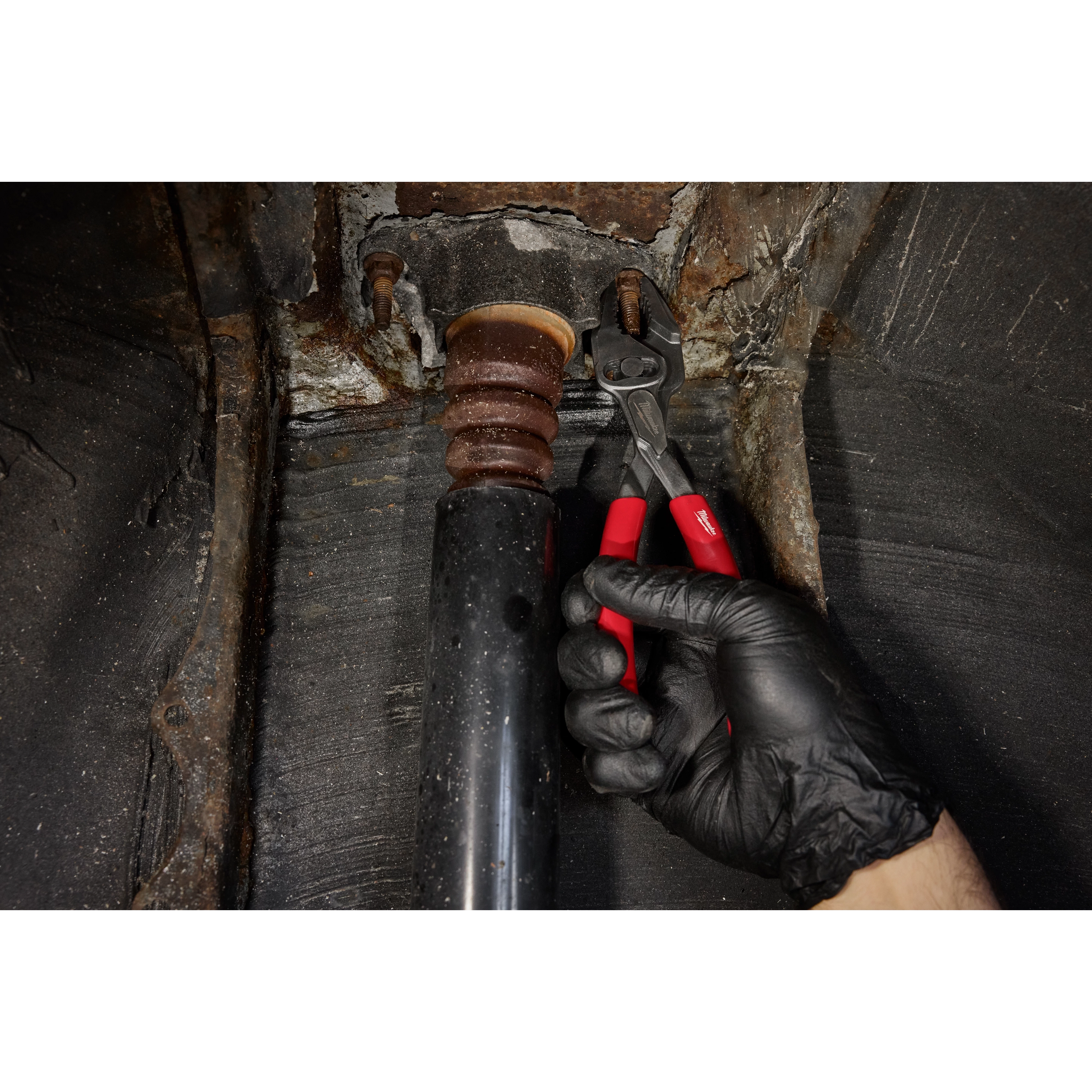 A hand wearing a black glove grips the 8" Slip Joint Pliers to adjust a metal object inside a black pipe. The pliers have a silver head and red handles. The background shows a rusty, textured surface.